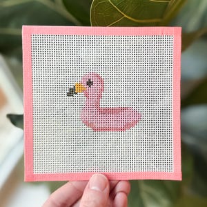 May include: A pink needlepoint canvas featuring a flamingo design. The flamingo has a black and yellow beak and is set against a white background. The canvas is framed with a pink border. The image is held by a hand.
