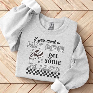 Könnte beinhalten: Grauer Pullover mit einem grafischen Design, auf dem "Wenn du ein Softeis willst, hol dir Eis" steht. Die Grafik enthält ein stilisiertes Eiswaffelhörnchen und ein kariertes Muster.