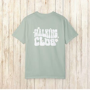 May include: A light green t-shirt with a white graphic that says "WALKING CLUB" in a retro font with small white flowers on either side of the text.