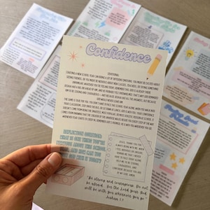 May include: A rectangular devotional card with a light cream background featuring the title Confidence in purple text. The card includes a prayer, a reflection question, and the Bible verse Joshua 1:9, decorated with illustrations of a pencil and sharpener.