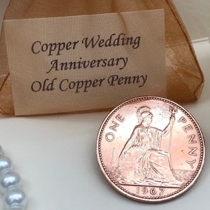May include: A copper penny from 1967 with the inscription "ONE PENNY" and the image of a woman holding a trident. The penny is in a small, brown, mesh bag with a tag that reads "Copper Wedding Anniversary Old Copper Penny".