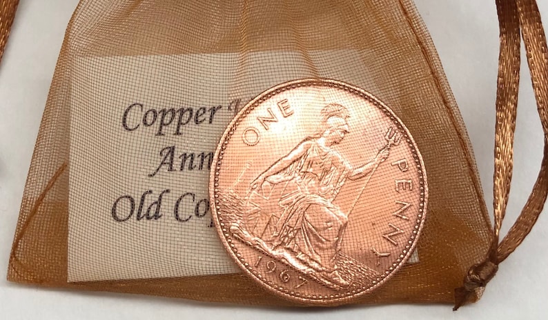 May include: A copper-colored 1967 British one penny coin with the inscription "ONE PENNY" and a depiction of Britannia. The coin is in a small, brown, mesh bag with a tag that reads "Copper 1st Anniversary Old Coin".