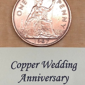 May include: A copper penny coin from 1967 with the inscription "ONE PENNY" and a depiction of Britannia. The coin is on a light brown surface with the text "Copper Wedding Anniversary Old Copper Penny".