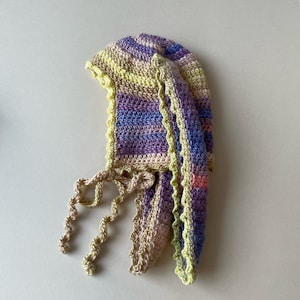 May include: A crocheted baby bonnet with a striped pattern in shades of purple, yellow, and pink. The bonnet has a scalloped edge and ties at the back.