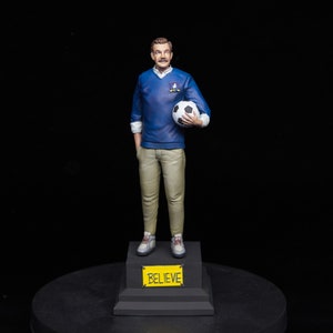 Könnte beinhalten: Figur einer Person in blauem Pullover und beiger Hose, die einen Fußball hält. Die Figur steht auf einem schwarzen Sockel mit einem gelben Schild "BELIEVE". Der Mann hat einen Schnurrbart und trägt weiße Turnschuhe.