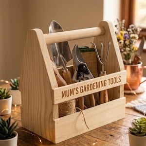 May include: A light-coloured wooden garden tool caddy with a handle, featuring the engraved text "MUM'S GARDENING TOOLS" on the front panel. The caddy contains a metal trowel, pruning shears, a small hand rake, and a roll of twine for gardening.