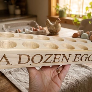May include: A rectangular light-coloured wooden egg holder featuring two rows of six circular indentations for a total of twelve eggs. The front side of the wooden block is engraved with the text A DOZEN EGGS in a dark, bold, serif font.