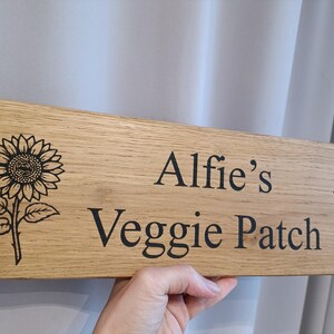 May include: Wooden sign with the text "Alfie's Veggie Patch" in black lettering. The sign features a sunflower illustration on each side. The wood has a natural grain.