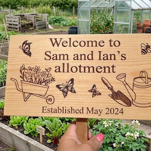 May include: A rectangular light-coloured wooden garden sign featuring laser-engraved text that reads Welcome to Sam and Ian's allotment and Established May 2024. The sign includes engraved illustrations of a wheelbarrow, butterflies, a bee, and a watering can.