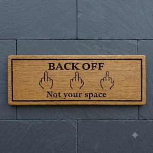 May include: A rectangular wooden sign with the words "BACK OFF" and "Not your space" engraved in a dark font. The sign also features three hand illustrations with the middle finger extended. The sign is mounted on a dark grey stone wall.