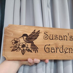 May include: A wooden sign with a hummingbird and flowers carved into it. The sign reads "Susan's Garden".