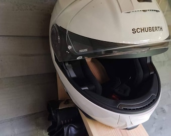 May include: A white motorcycle helmet with a clear visor and the word "SCHUBERTH" printed on the side. The helmet is mounted on a wooden shelf with a pair of black motorcycle gloves hanging below.