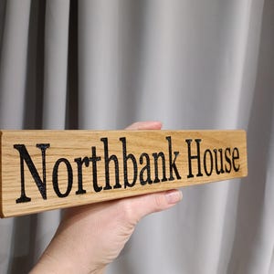 May include: A wooden sign with the words "Northbank House" in black lettering. The sign is rectangular and made of light-colored wood. The background is a plain grey curtain.