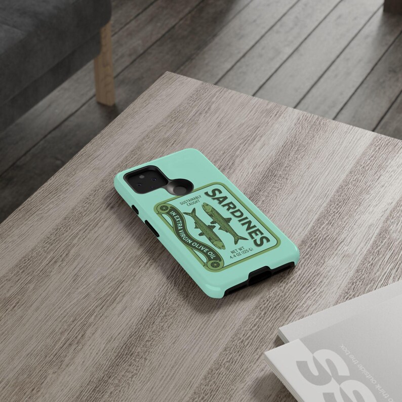 Peut inclure: Une coque de t&eacute;l&eacute;phone bleu turquoise avec un design de bo&icirc;te de sardines de style vintage. L'&eacute;tiquette de la bo&icirc;te indique "Sardines" en texte blanc avec un graphique d'huile d'olive verte et le texte "In Extra Virgin Olive Oil" et "Sustainably Caught".