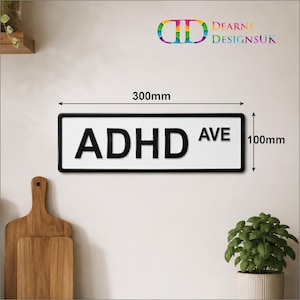 May include: A rectangular white sign with a black border and the black text ADHD AVE. The sign features dimension markers indicating a width of 300mm and a height of 100mm. The logo for Dearne DesignsUK is visible in the top right corner.