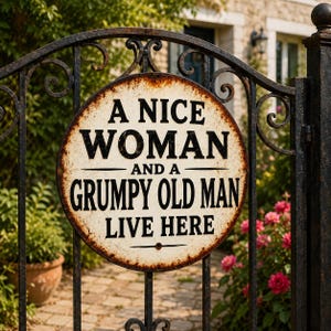 May include: A circular metal sign with a distressed, rusted edge featuring the black text A NICE WOMAN AND A GRUMPY OLD MAN LIVE HERE. The sign is mounted on a black wrought iron gate, displaying a weathered, vintage-inspired finish throughout.