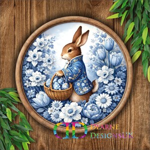 May include: A circular print with a brown rabbit in a blue coat, holding a basket of blue Easter eggs, surrounded by blue and white flowers. The print is set in a wooden frame against a wooden background with green leaves.