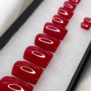 Puede incluir: Un conjunto de diez uñas postizas rojas de diferentes tamaños. Las uñas están dispuestas en una fila en una caja negra con un interior blanco.