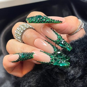 May include: Long, almond-shaped artificial nails with a French manicure design. The tips are adorned with emerald green rhinestones. A large, silver ring with clear stones is on the ring finger. Another silver ring is on the index finger.