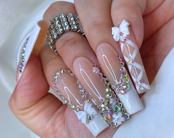 May include: Long, almond-shaped nails with a white base and intricate crystal and bow designs. The nails are decorated with white bows, clear crystals, and white lines.