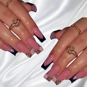 Puede incluir: Primer plano de manos con uñas acrílicas largas, cuadradas, de color rosa, con puntas negras y detalles de purpurina dorada y flores. Las uñas están decoradas con pequeños abalorios dorados en forma de flor. El texto "ZAMI D'OR BEAUTY" es visible en la mano izquierda.