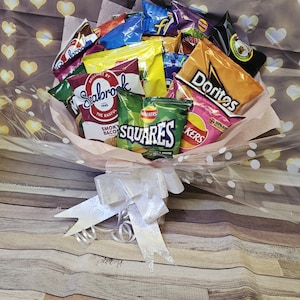 May include: A bouquet of various chip bags, including Doritos, Walkers Squares, and Seabrook, arranged in a clear plastic wrap with white polka dots and a white satin bow. The bouquet is sitting on a wooden surface.