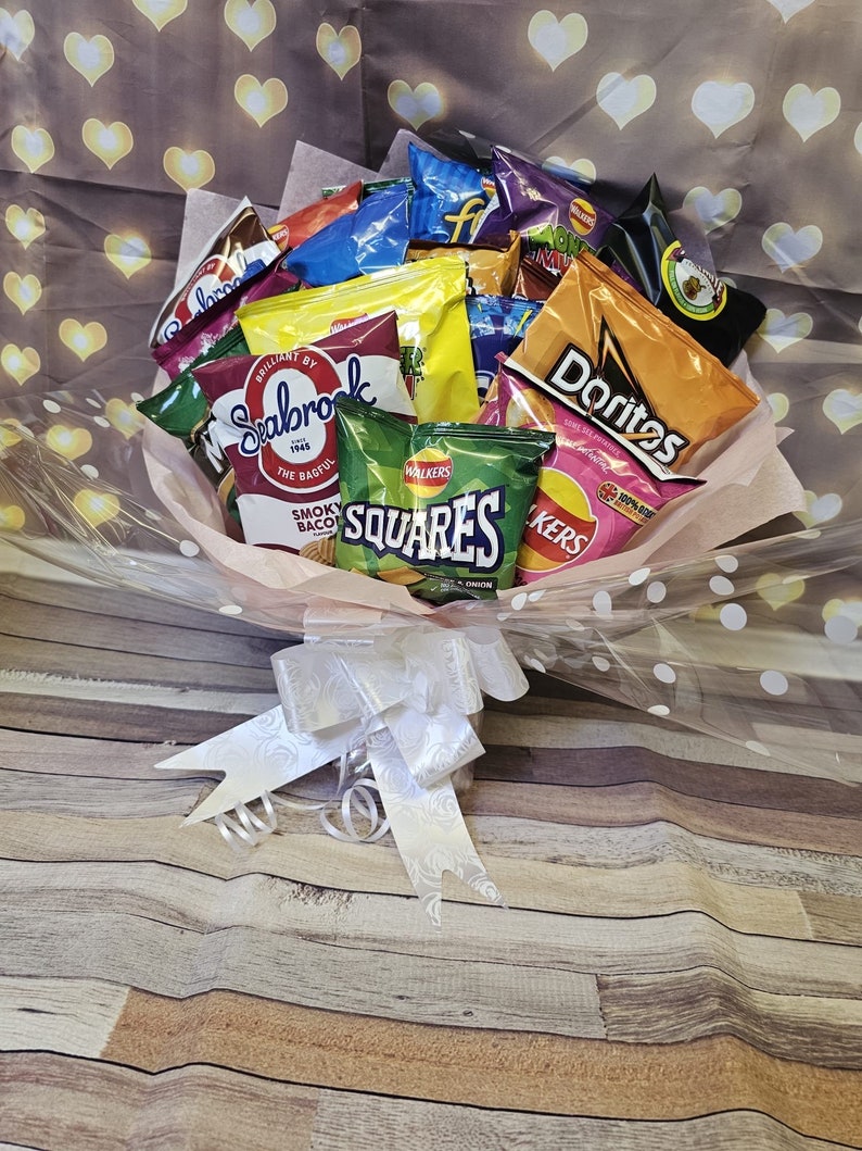 May include: A bouquet of various chip bags, including Doritos, Walkers Squares, and Seabrook, arranged in a clear plastic wrap with white polka dots and a white satin bow. The bouquet is sitting on a wooden surface.