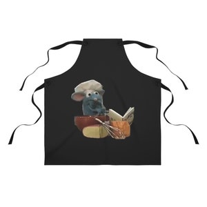 May include: Black apron with a cartoon rat chef wearing a white chef's hat, holding a cookbook and whisk. The rat is in front of a red bowl with cheese and other ingredients. The apron has black ties.