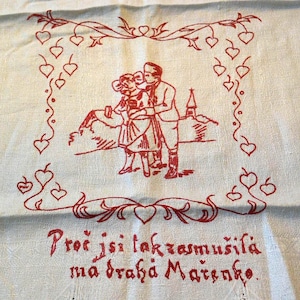 May include: Cream-colored textile with red embroidered design. The design features a couple embracing, a church, and a border of hearts. Text in red script is at the bottom. The textile is likely a decorative piece.