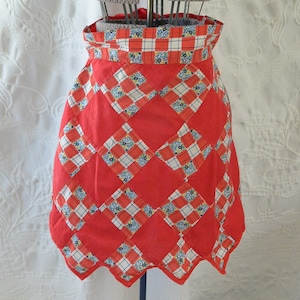 May include: A red quilted apron with a white and blue checkered pattern. The apron has a scalloped hem and is made of cotton fabric.
