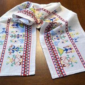 May include: A white waffle-weave kitchen towel featuring colorful cross-stitch embroidery. The design includes repeating patterns of stylized figures, floral motifs, and geometric borders in shades of red, blue, yellow, and green thread.