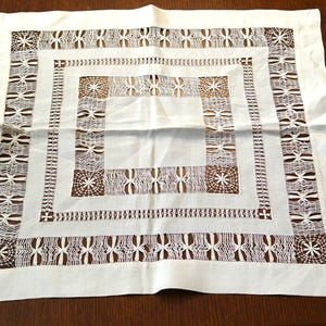 May include: A square, off-white linen tablecloth with an intricate, openwork design. The design features a series of concentric squares with floral and geometric patterns. The tablecloth is placed on a dark brown wooden surface.