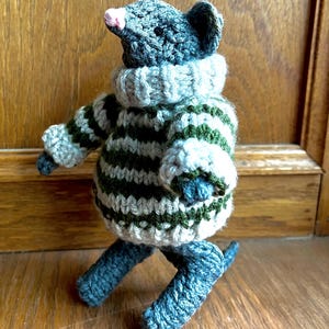 May include: A handmade crochet mouse figurine. The mouse is gray with a pink nose and wears a green and white striped sweater and a blue turtleneck. The mouse has blue legs and a tail.