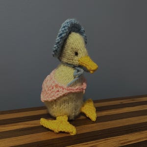 May include: A handmade knitted duckling wearing a light blue bonnet and a pink shawl. The duckling has a yellow beak and feet. The toy is sitting on a wooden surface with brown and tan stripes.