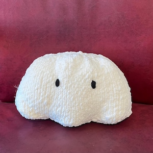 May include: A white, cloud-shaped decorative cushion with two black dot eyes. The cushion has a soft, textured appearance and is placed on a burgundy surface. This plush cushion adds a whimsical touch to any room.