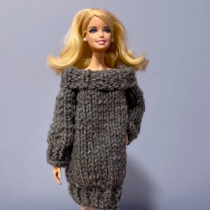 Könnte beinhalten: Eine Puppe in einem grauen, schulterfreien Strickkleid. Das Kleid hat lange Ärmel und einen grob strukturierten Strick. Die Puppe hat blondes Haar und kleine weiße Ohrringe. Der Hintergrund ist einfarbig hellblau.