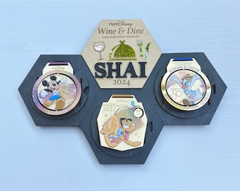 Può includere: Tre medaglie d'oro con nastri neri, esposte su un espositore esagonale in legno. Le medaglie raffigurano Topolino, Pluto e Paperino, e commemorano il 50° anniversario di Walt Disney World. L'espositore include anche una targa in legno con il testo "runDisney Wine & Dine Half Marathon Weekend SHAI 2024".