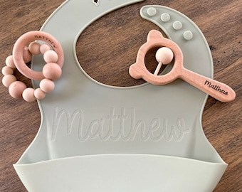 Puede incluir: Un babero de silicona verde salvia con el nombre "Matthew" grabado en él. El babero se acompaña de dos mordedores de madera, uno con un anillo de cuentas y el otro con forma de oso con un sonajero.