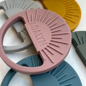 May include: A set of four silicone teethers in the shape of a sun with a textured surface. The teethers are in the colors gray, mustard yellow, dusty rose, and teal. The dusty rose teether has the word "ADALY" embossed on it.