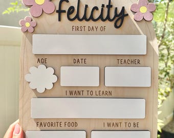 May include: Wooden first day of school sign for Felicity, with pink and yellow flower accents. The sign includes spaces to write age, date, teacher, favorite food, and what the child wants to learn and be.