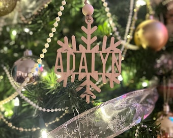 Op de afbeelding: Een houten sneeuwvlok kerstversiering met de naam "ADALYN" uitgesneden in het midden. De versiering hangt aan een touwtje en hangt voor een versierde kerstboom met gouden en zilveren ballen.