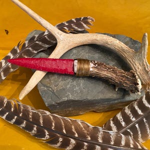 May include: A handcrafted knife with a red blade and a deer antler handle, wrapped with twine. The knife rests on a gray stone, with feathers and a deer antler in the background. The background is a solid yellow color.