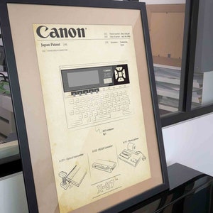 以下が含まれることがあります： キヤノンX-07ハンドヘルドコンピューターのヴィンテージな白黒イラスト。イラストには、「キヤノン」、「日本特許」、「5461」、「ハンドヘルドコンピューター」、「X-721」、「光学トランスミッター」、「X-722」、「RS232Cコンバーター」、「X-711」、「サーマルプリンター」、「X-07」のテキストが含まれています。