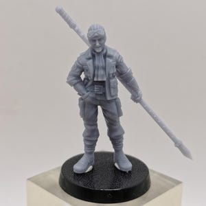 May include: A gray plastic miniature figurine of a person wearing a jacket, pants, and boots, holding a long spear. The figurine is standing on a black circular base.