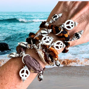 May include: A collection of bohemian-style bracelets. Each bracelet features a white peace sign charm, decorative beads, and fabric accents in shades of brown, black, and cream. The bracelets are displayed against a backdrop of a beach scene.