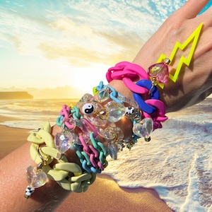 May include: A collection of colorful bracelets adorns a wrist, featuring a variety of styles and materials. The bracelets include chunky chain links in pastel shades, clear beaded strands, and charms. A neon yellow lightning bolt charm adds a pop of contrast.