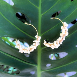 May include: A pair of gold-toned hoop earrings adorned with small, irregularly shaped, off-white stones. The earrings are set against a large, green leaf with several holes, creating a natural backdrop.