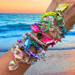 May include: A collection of colorful bracelets with various charms, including hearts, stars, and lightning bolts. The bracelets feature a mix of clear, neon, and iridescent elements, creating a vibrant and playful aesthetic. The background shows a beach scene with the ocean and sunset.