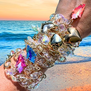Puede incluir: Una colección de pulseras doradas y transparentes adornadas con dijes de corazón, hoja y piedras preciosas. Las pulseras presentan una variedad de cristales transparentes, iridiscentes y coloridos. La imagen está ambientada en una playa con una puesta de sol.