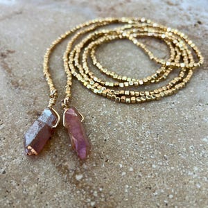 May include: A long necklace featuring a strand of small, metallic gold-colored square beads. The necklace ends in two dangling, raw-cut crystal points with a translucent pink hue, each secured with gold-colored wire wrapping at the top.